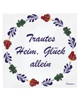 A square white ceramic tile featuring the German text 'Trautes Heim, Glück allein' in dark purple cursive script, framed by a hand-painted traditional Dutch floral border in red, green, and purple with small gold accents Boerenbont style. [market:de, at]