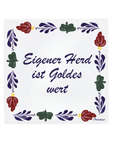 A square white ceramic tile featuring the German text 'Eigener Herd ist Goldes wert' in dark purple cursive script, framed by a hand-painted traditional Dutch floral border in red, green, and purple with small gold accents Boerenbont style. [market:de, at]
