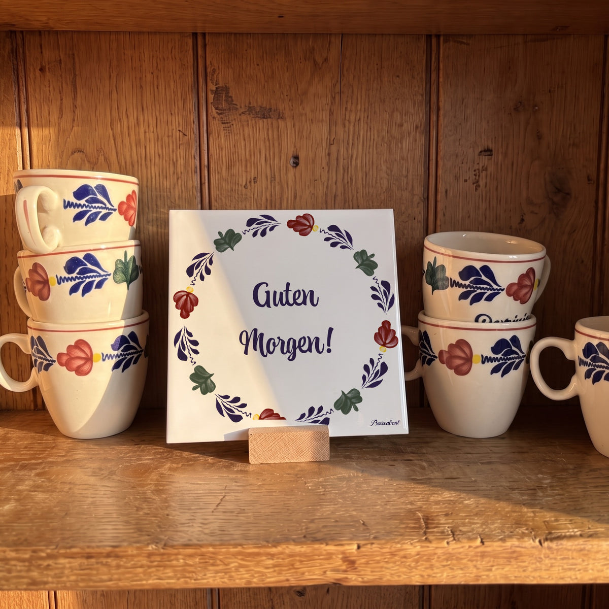 Boerenbont ceramic tile featuring the German greeting "Guten Morgen!" surrounded by traditional floral patterns, displayed with matching coffee mugs. [market:de,at]