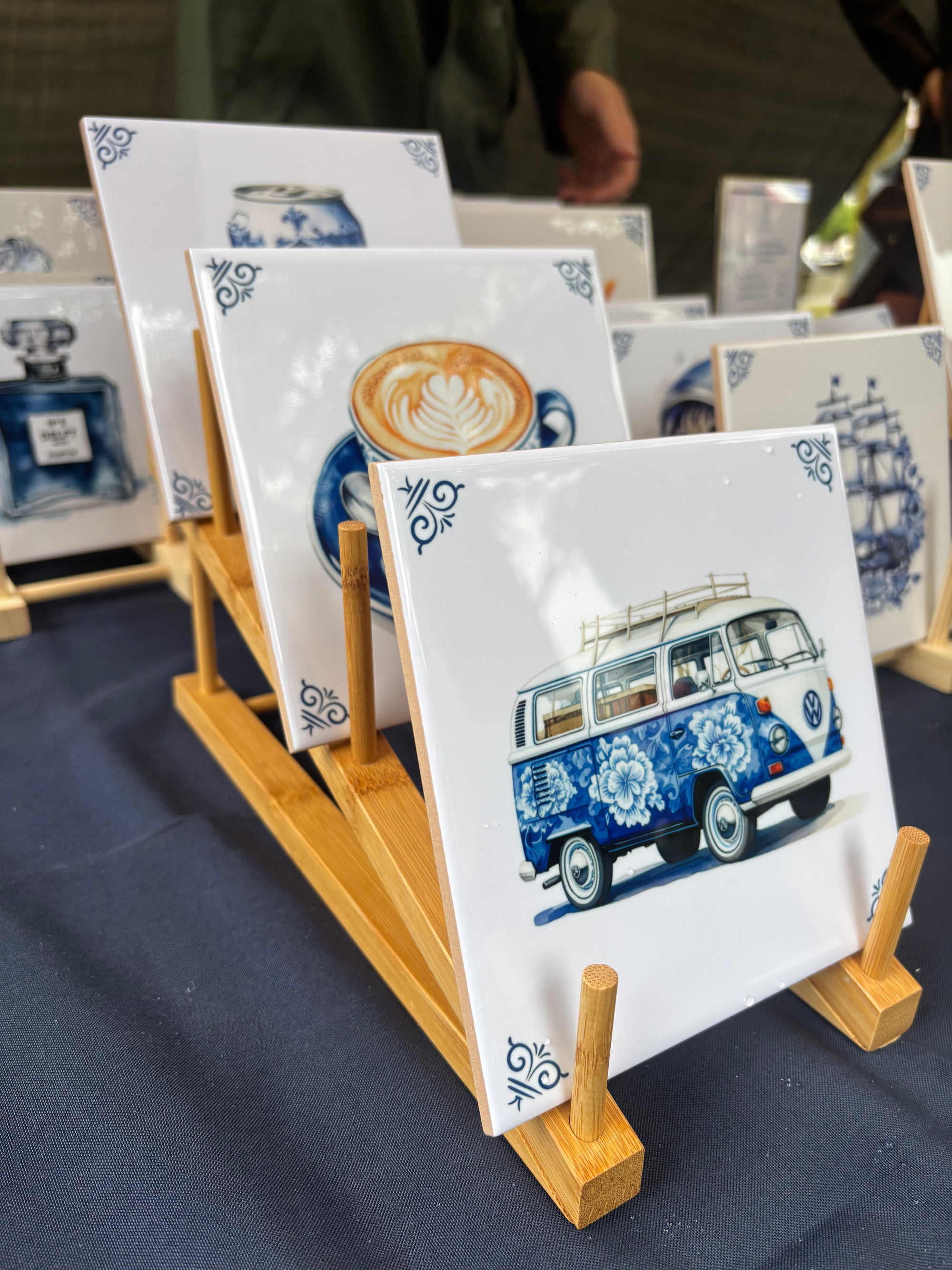 A set of square white ceramic tiles with blue and multicolor illustrations, displayed on small wooden stands on a dark tablecloth. The front tile features a blue and white patterned VW bus, while other visible tiles show a coffee cup with latte art, a perfume bottle, and a sailing ship.