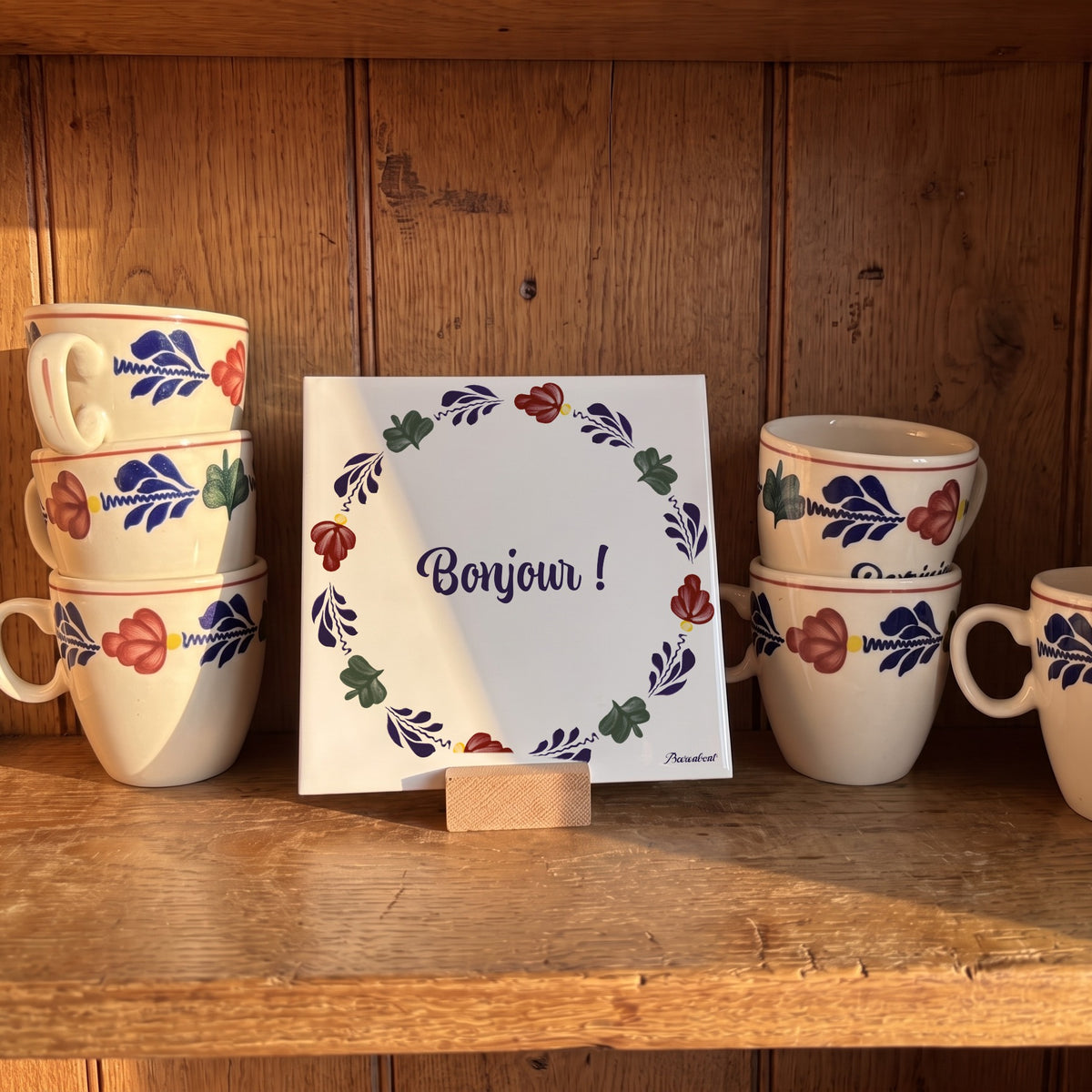 Decorative Boerenbont ceramic tile featuring the French greeting "Bonjour!" framed by traditional floral motifs, displayed on a wooden shelf alongside matching Boerenbont coffee cups. [market:fr]