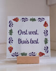 Traditional Boerenbont ceramic tile featuring the Dutch saying "Oost west, thuis best" surrounded by colourful floral motifs on a wooden tile stand.