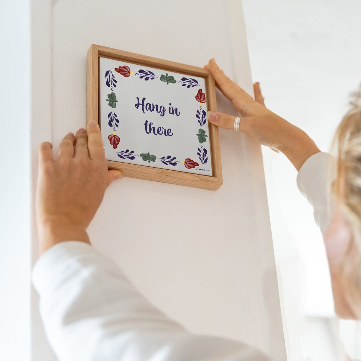 Boerenbont ceramic tile featuring the motivational quote "Hang in there" and traditional floral patterns, displayed in a wooden frame on a white wall.