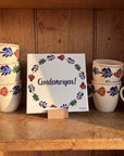 Decorative Boerenbont ceramic tile featuring the Dutch greeting "Goedemorgen!" surrounded by colourful traditional floral patterns, displayed alongside matching Boerenbont coffee mugs. [market:nl]