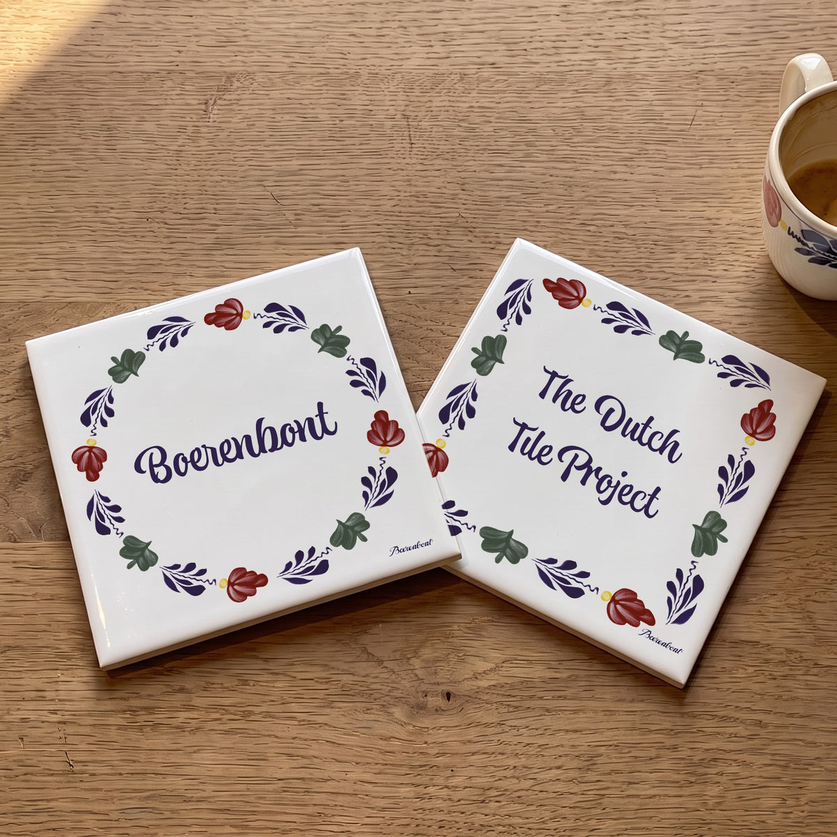 Two decorative ceramic tiles featuring the Boerenbont and The Dutch Tile Project logos, surrounded by traditional Dutch floral patterns, displayed on a wooden table.