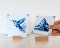 Two Delft Blue tiles displayed with wooden tile holders, one held by hand above the stand