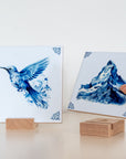 Two Delft Blue tiles displayed with wooden tile holders, one held by hand above the stand