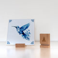 Delft Blue hummingbird tile displayed in a wooden holder next to a branded wooden tile stand