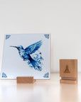 Delft Blue hummingbird tile displayed in a wooden holder next to a branded wooden tile stand