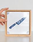 Wooden frame with a 15x15 cm chef’s knife tile inside, being placed upright on a table by someone’s hand.