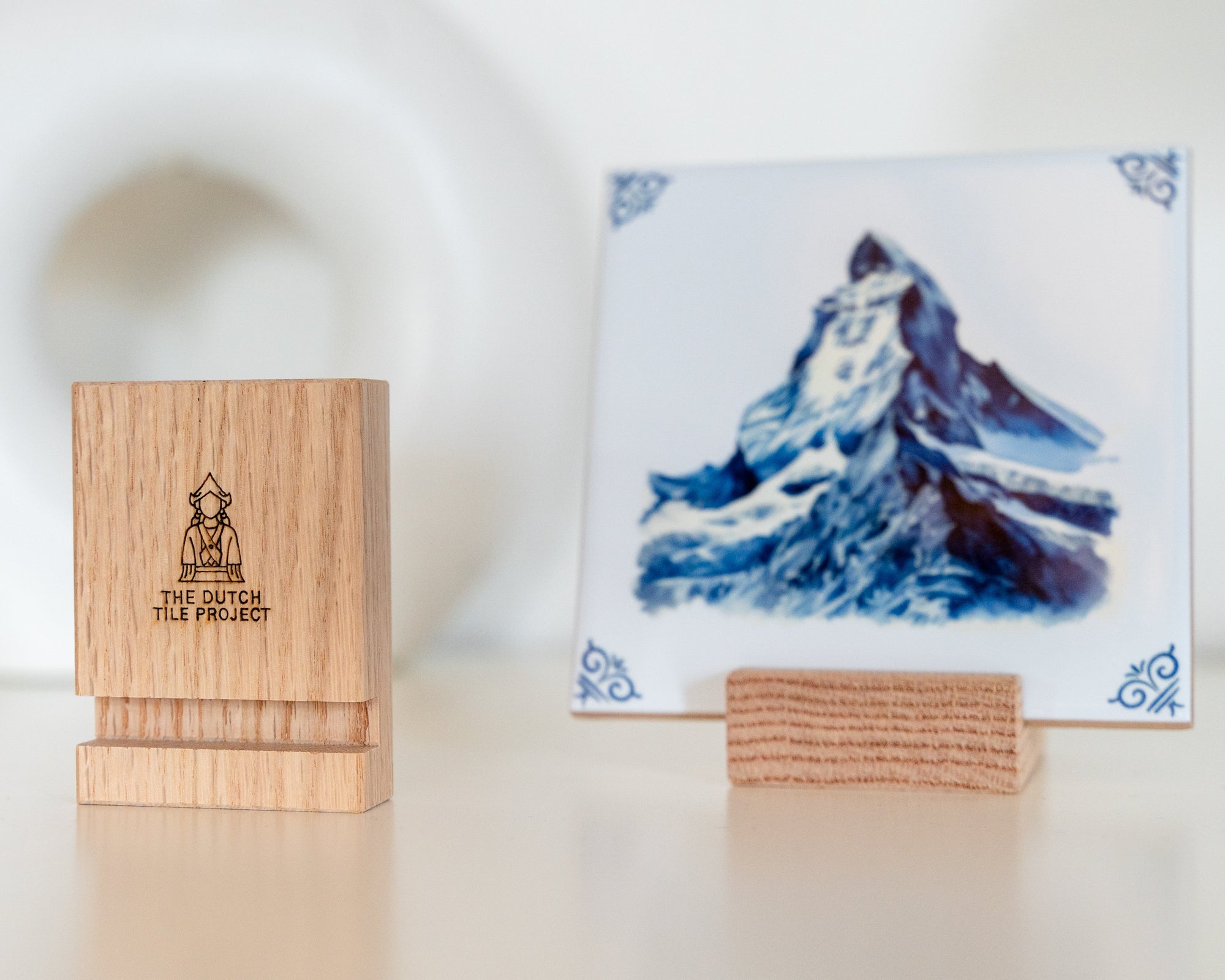 Delft Blue hummingbird tile displayed in a wooden tile holder next to a second wooden holder with The Dutch Tile Project logo